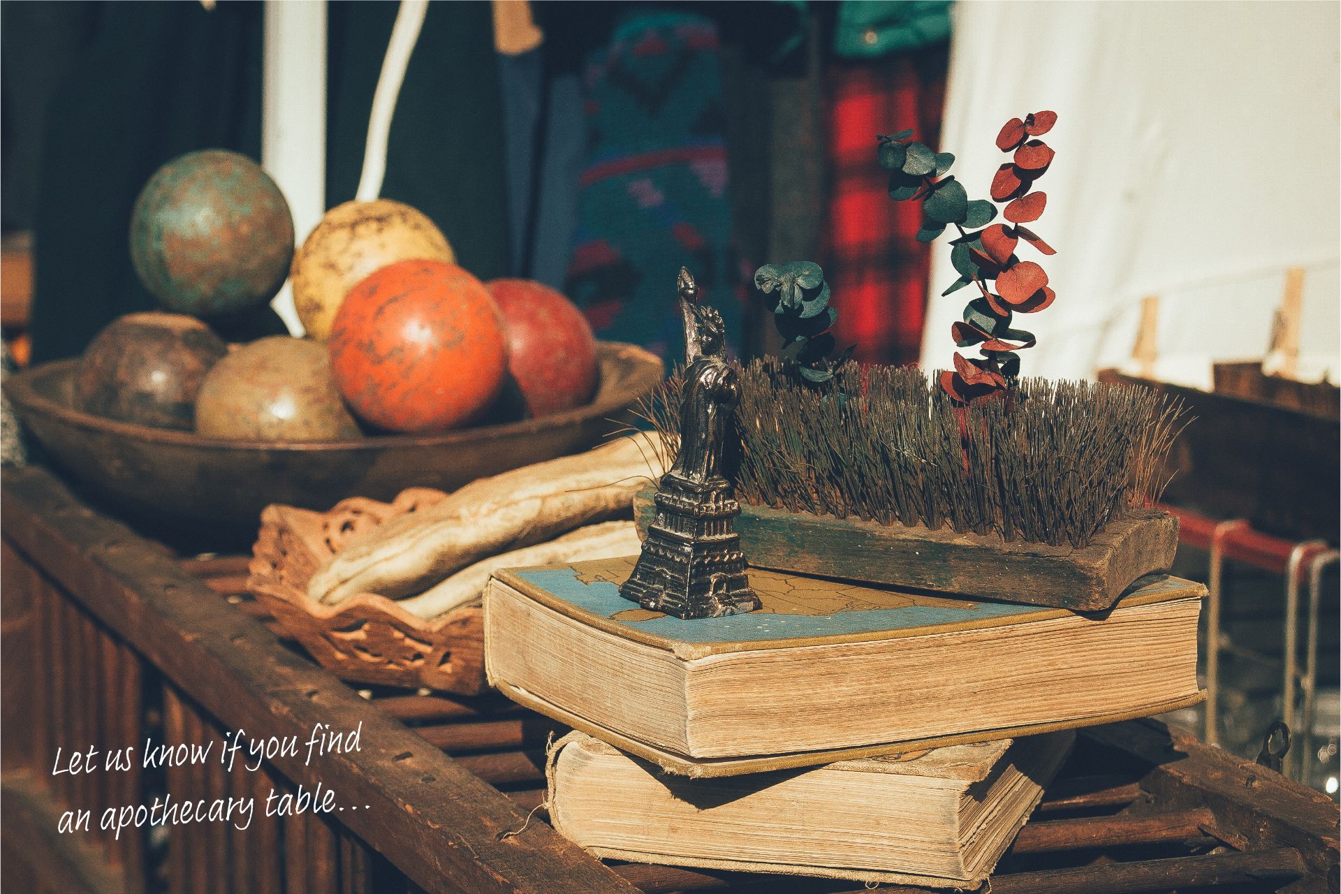 Table at a flea market in New York full of old books and bric-a-brac, with a handwritten piece of text saying, "Let us know if you find an apothecary table..."