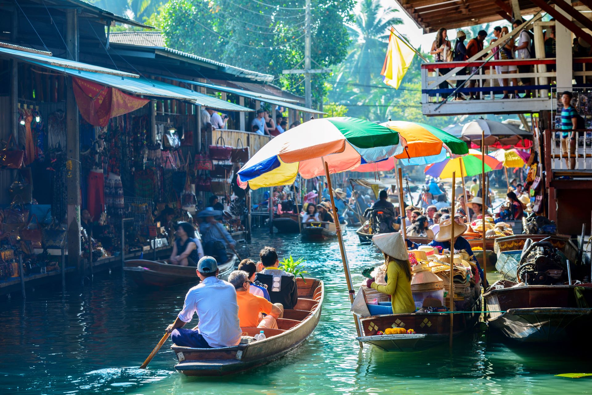 Three people sail on a boat through a bustling floating market in Bangkok, Thailand.