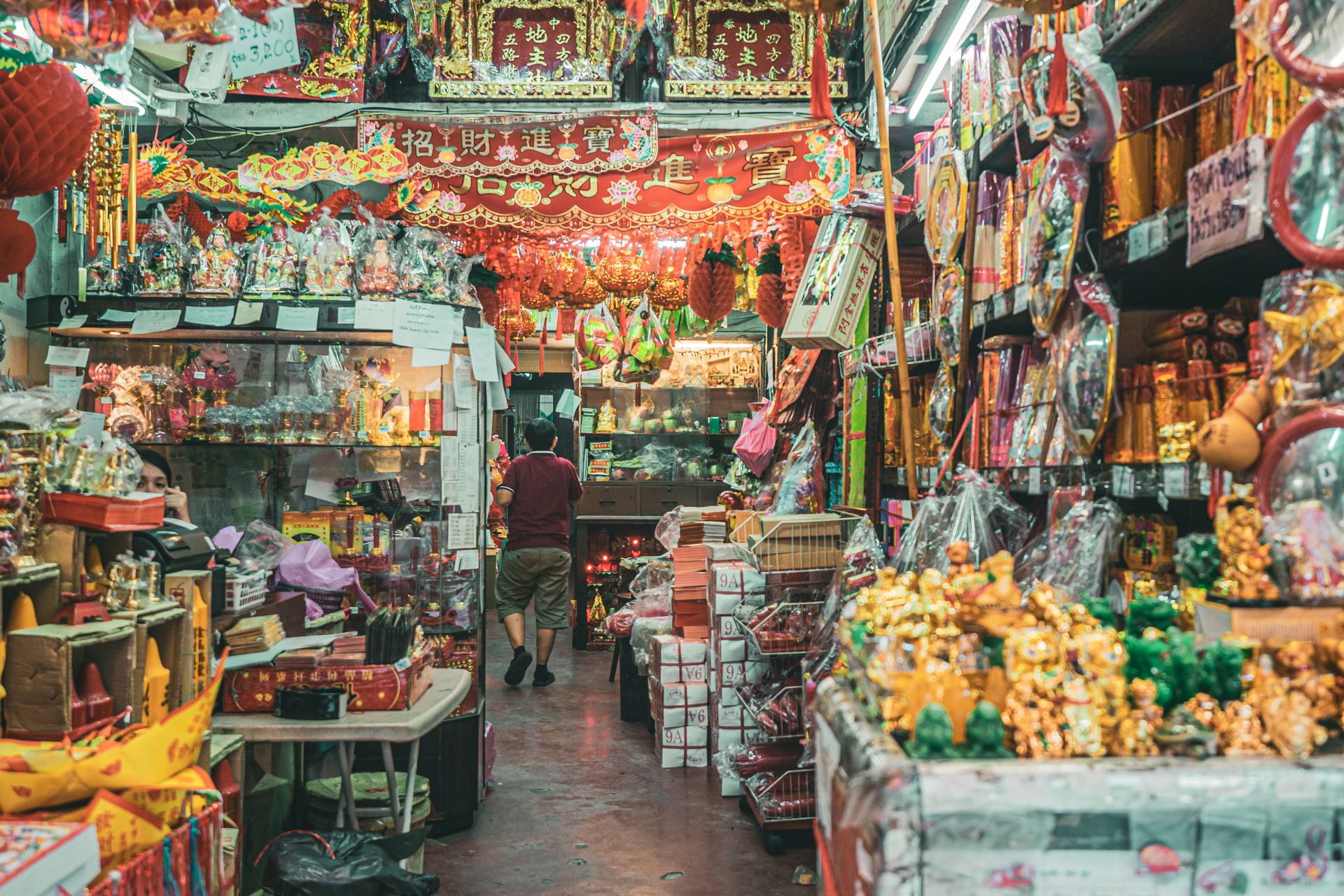 A colourful indoor market scene with tables and shelves full of haphazard produce and a man walking away in the background.