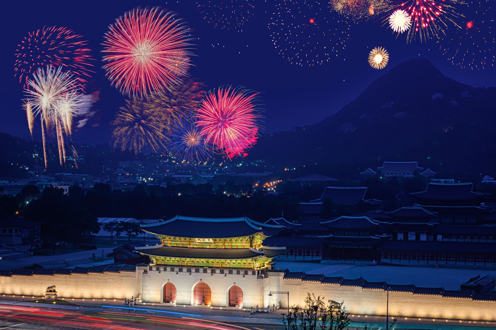 A Korean temple is lit up by fireworks in the sky above, whilst mountains loom in the background.