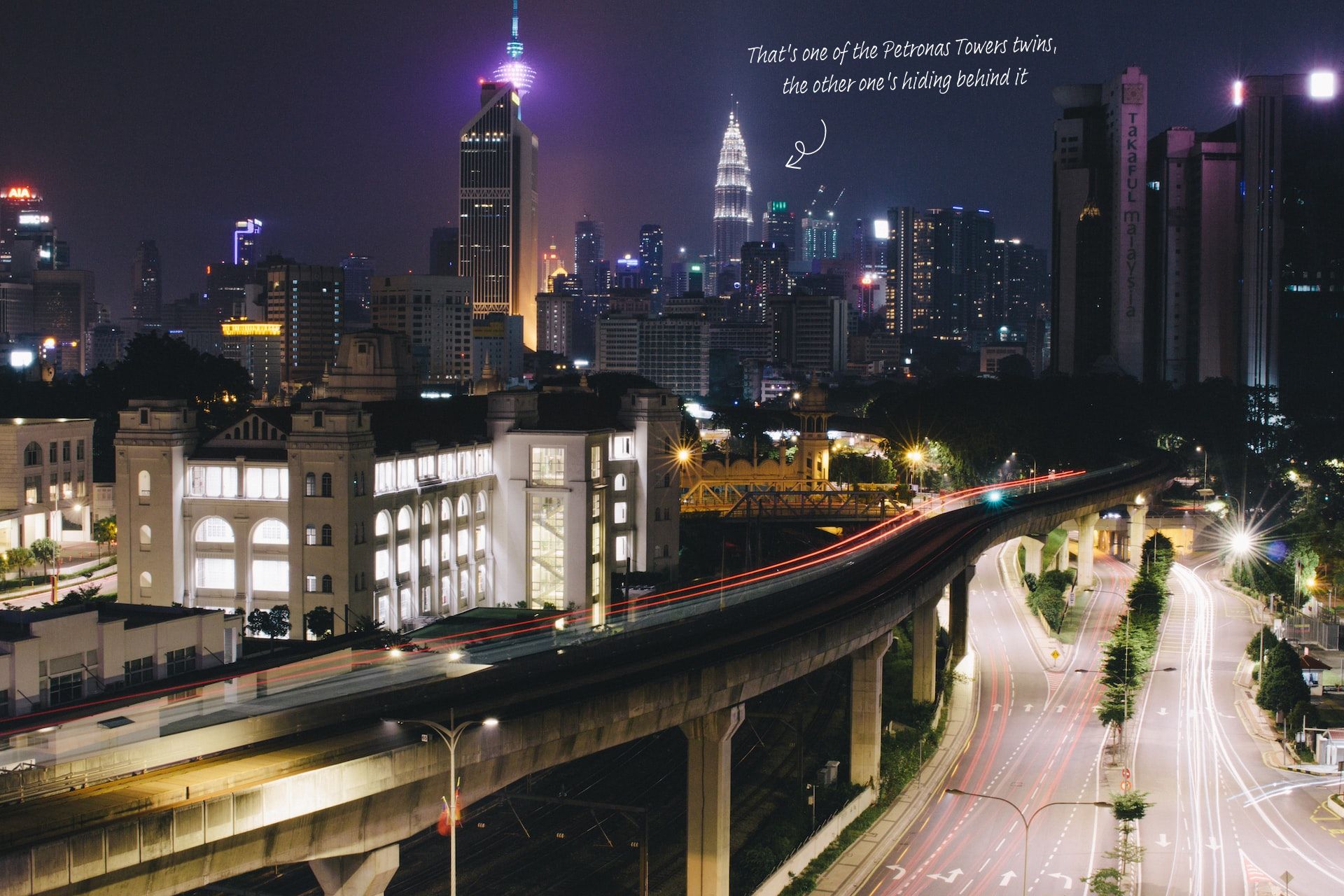 The skyline of Kuala Lumpur at night, with the KL Monorail in the foreground and the Petronas Towers glowing in the far.
