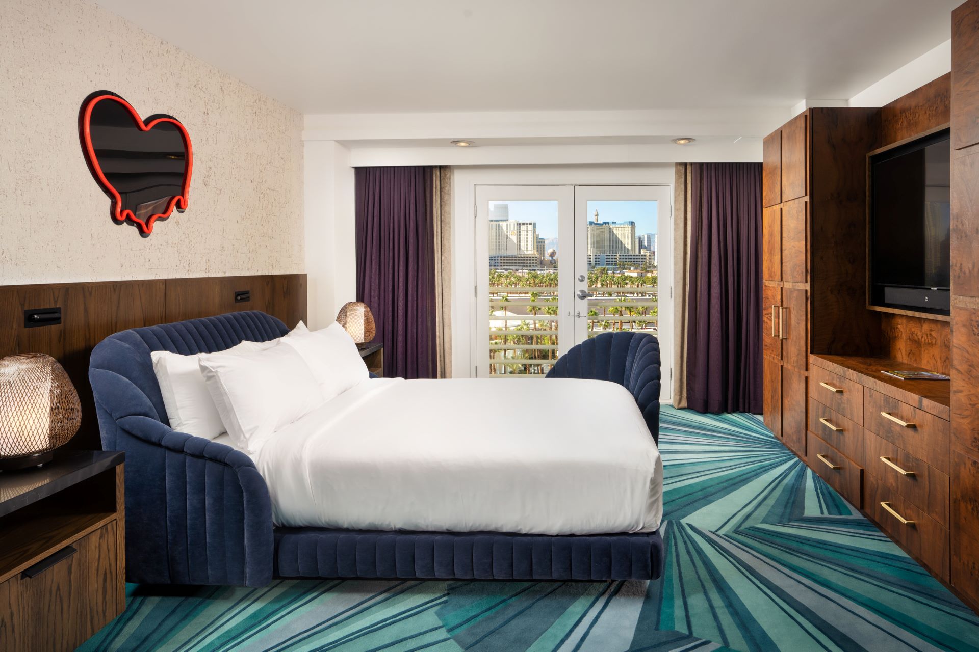 The second bedroom in the Shag Suite at Virgin Hotels Las Vegas. A blue, abstract pattern covers the carpet, a large bed in the cntre of the room with a kitschy neon heart sign above the headboard.