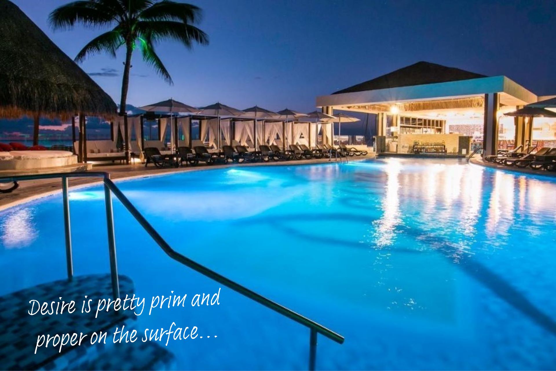 The pool at Desire Resort Cancun. Ground level view of the pool with bright blue water, surrounded by palm trees and deck chairs. Evening shot.