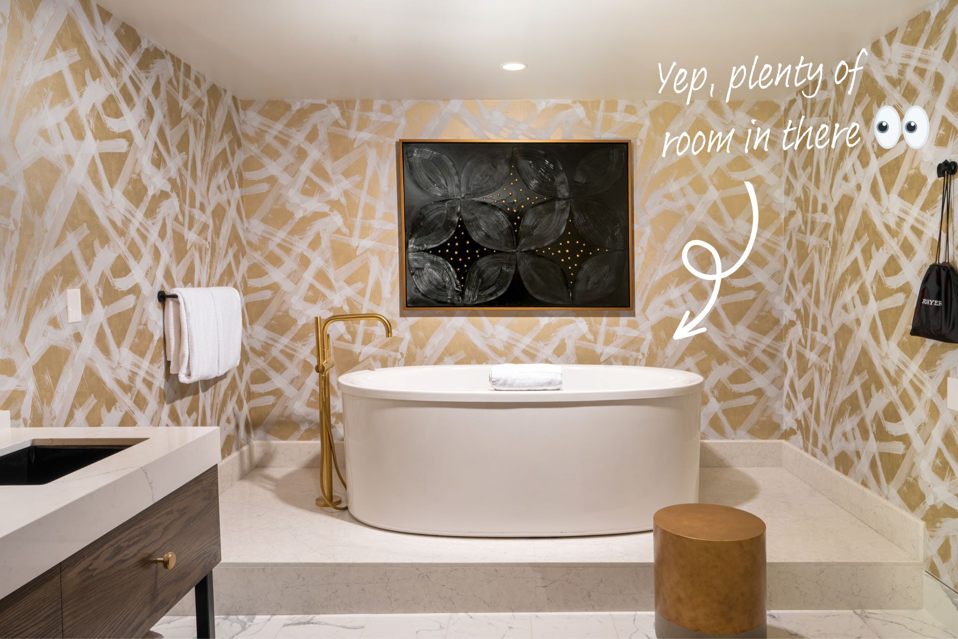 The bathroom in the Shag Suite at Virgin Hotels Las Vegas. A modern stand alone bath sits in the centre of the room, a large abstract painting above it, on a wall decorated with yellow patterned wallpaper.