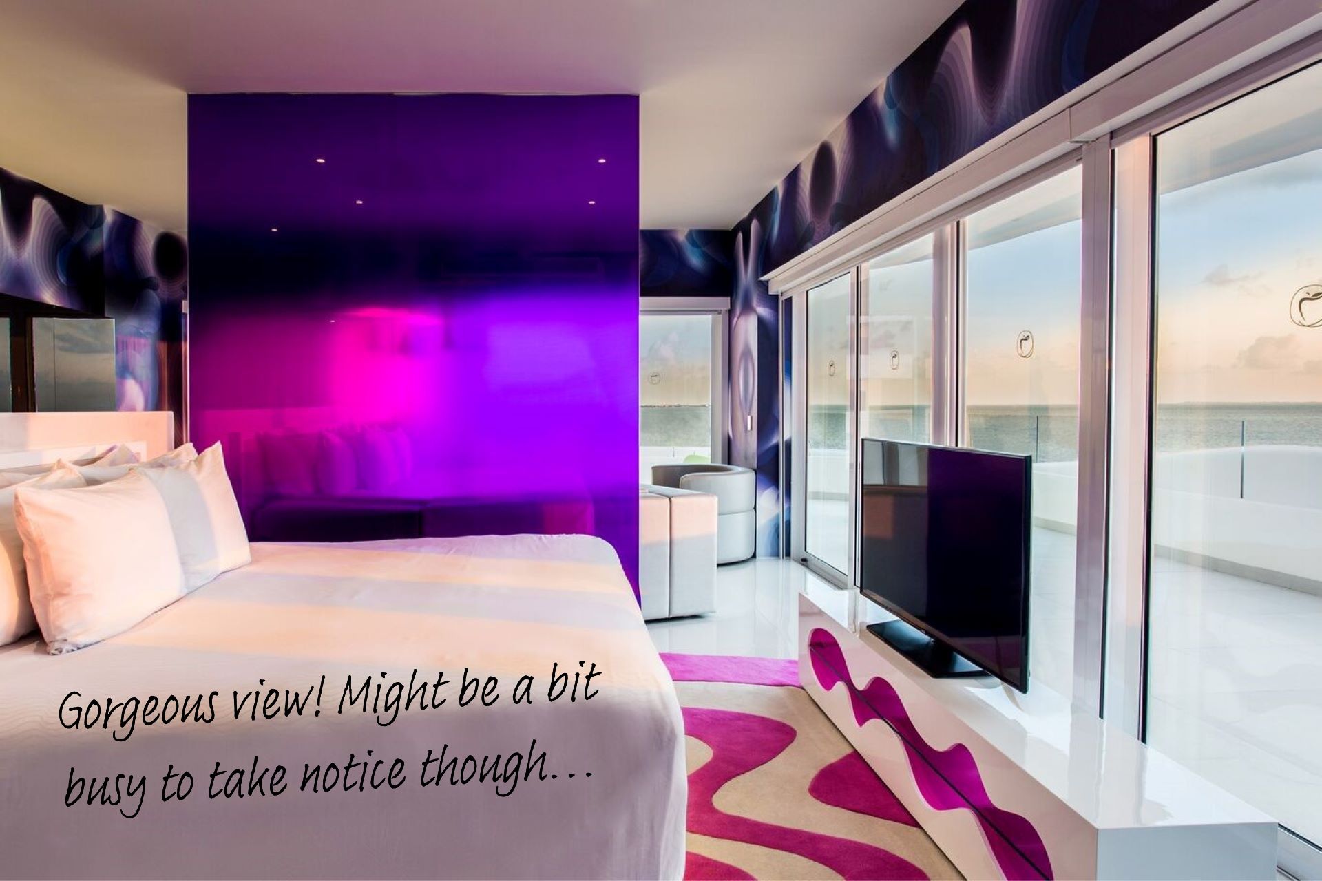 The Ocean Front Master Suite at Temptations Resort Cancun. A large white bed faces a panoramic view of the ocean, with a purple partitioning wall covered in a reflective material next to it.