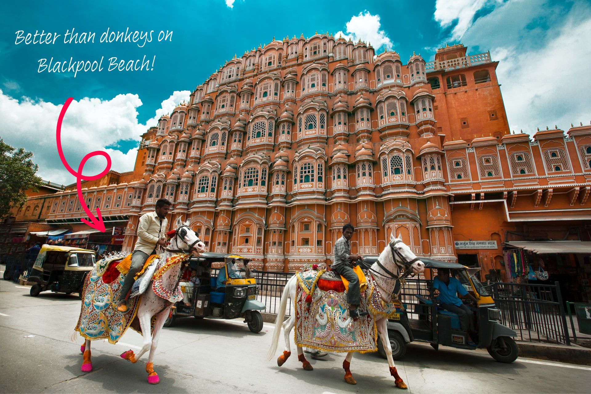 A low angle shot of a colonial style building in Rajasthan, India. Two men ride horses that have been decorated with colourful shawls.