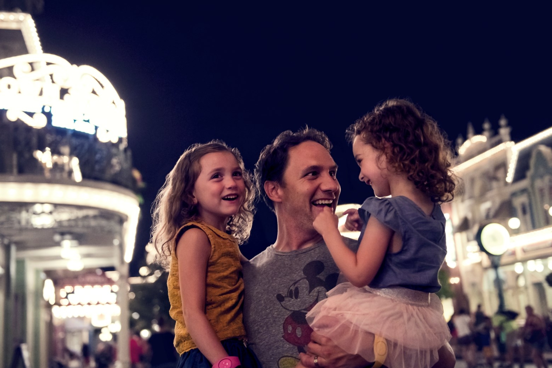 A father holds his two daughters and smiles, Magic Kingdom park at Disney in the background. Night.