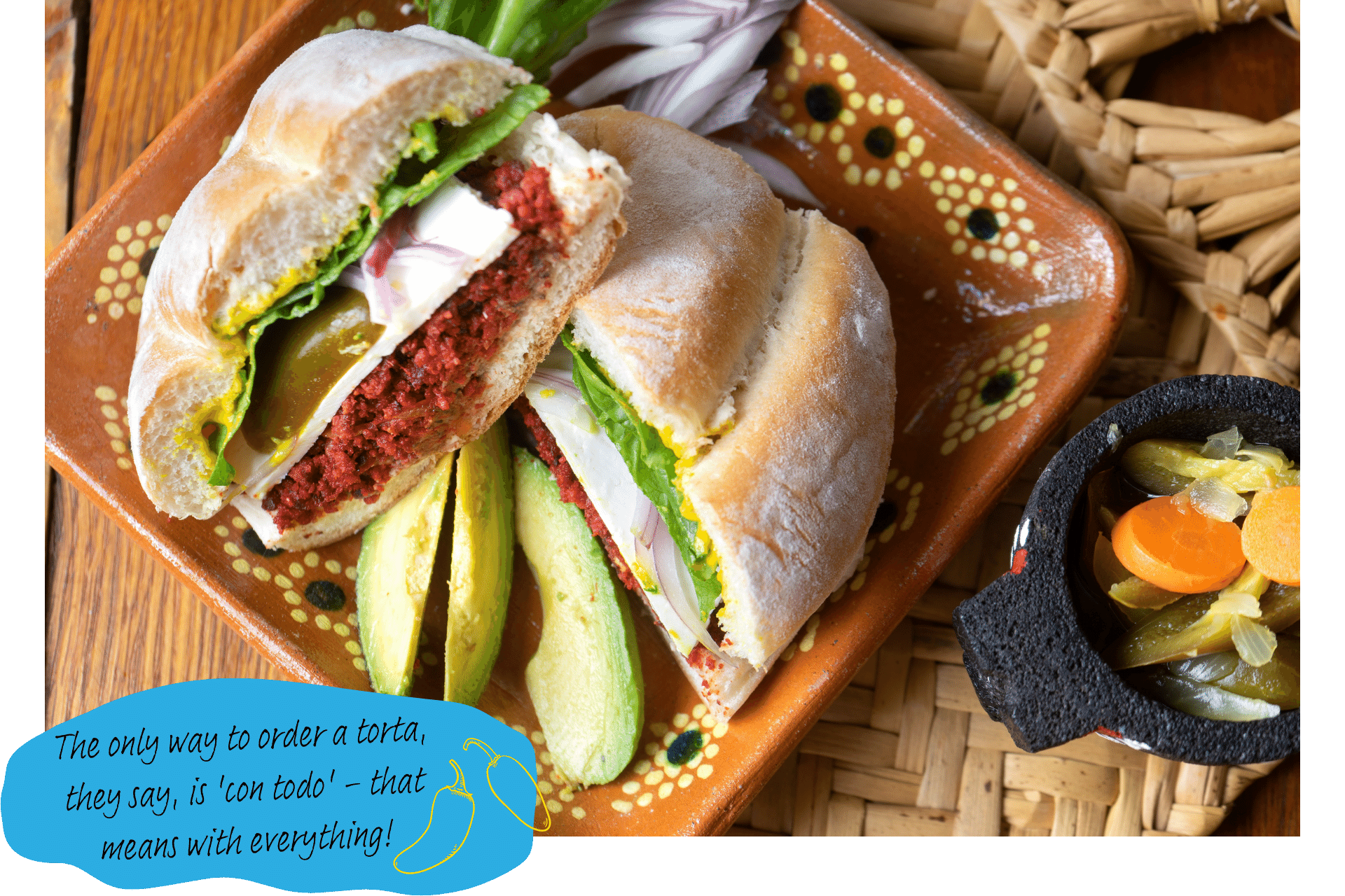 A Mexican torta sandwich sits on a plate which is decorated with various ingredients used in it, including onions and avocado.