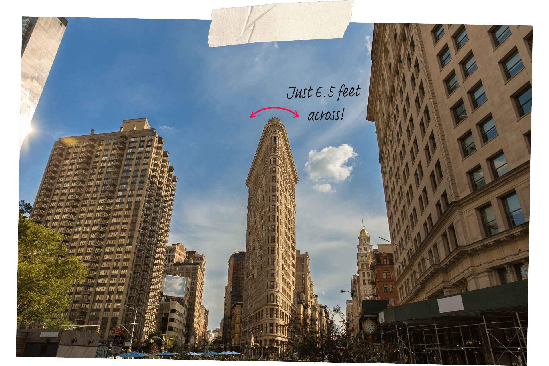 The Flat Iron Building is one of the must-visit historic sites in NYC. Image shows the narrow front facade of the Flat Iron Building from ground level, mid-rise building on either side of it.