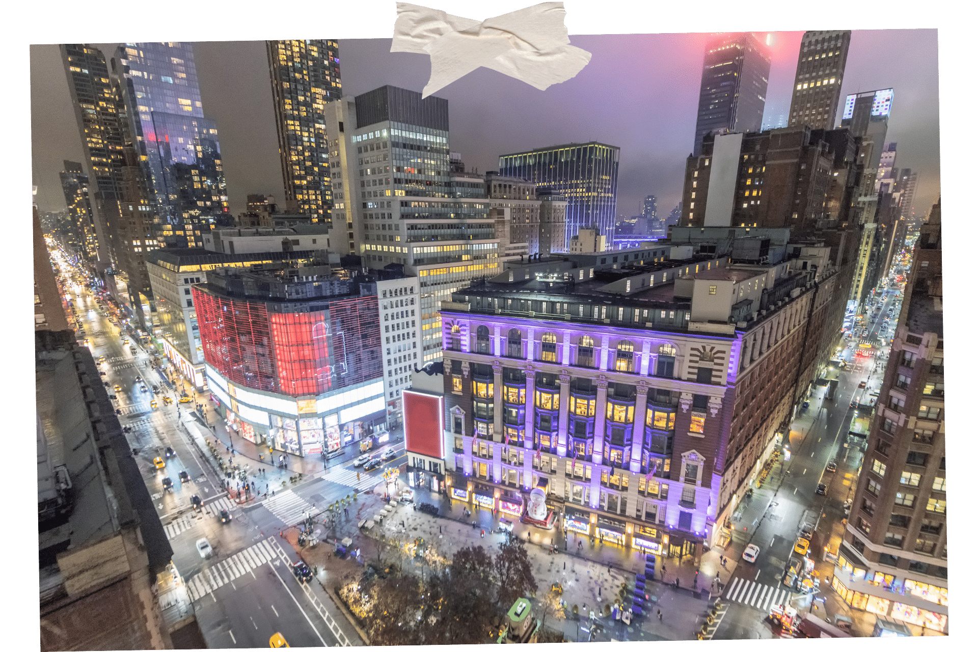 Herald Square is full of NYC's must-visit stores. Image shows a busy shopping area at night from above. Midrise buildings are lit up in various colours, skyscrapers loom all around, and shoppers pass by down on the ground below.